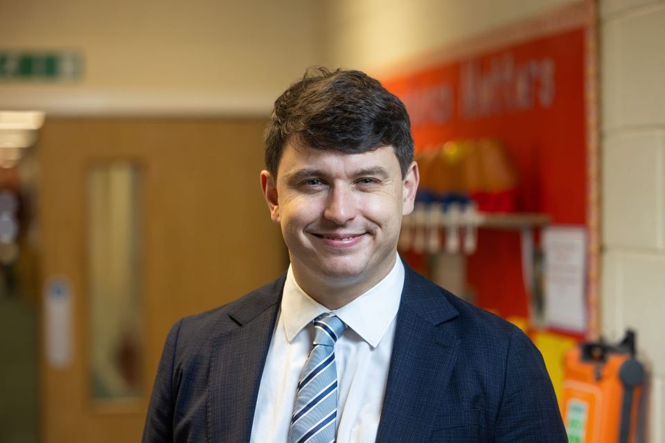 A man named Leon Tootle in a suit and tie is smiling in a hallway with a blurred background.